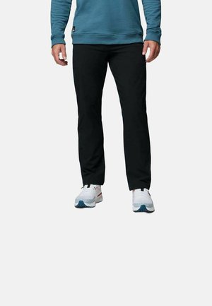 Mann trägt schwarze Hose, weiße Sneakers und ein blaues Langarmshirt und steht vor einem einfarbigen Hintergrund.