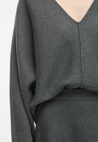 Sivkna pletenina s V-izrezom, ki ima ohlapen kroj, rebrast rob in gladko teksturo. Tkanina izgleda mehko in toplo.