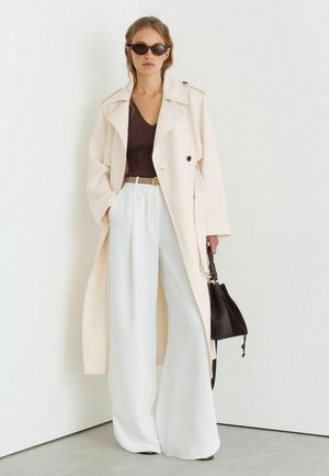 Frau trägt übergroßen cremefarbenen Trenchcoat, braunes Oberteil, weite weiße Hose, Sonnenbrille und hält eine schwarze Handtasche vor einer weißen Wand.