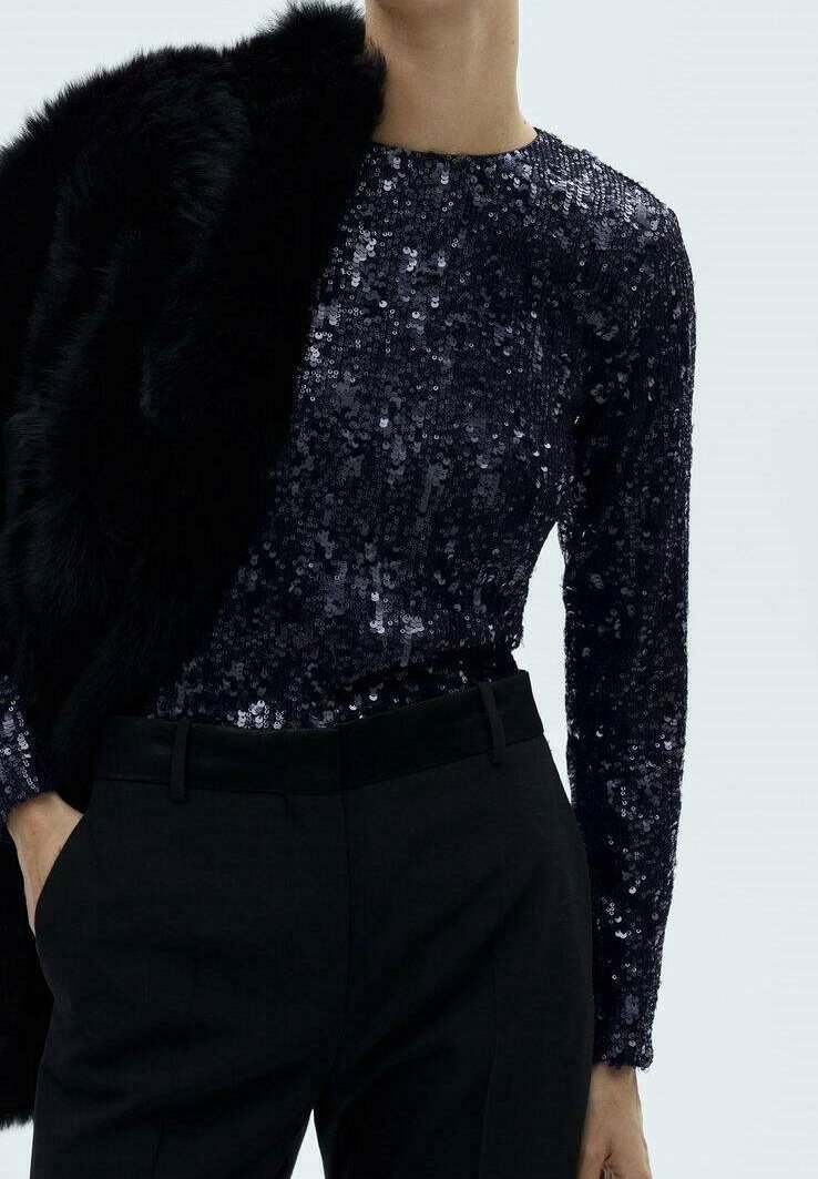 Personne portant un haut noir à manches longues à sequins et un pantalon noir, avec un manteau en fourrure noir drapé sur une épaule.