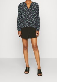 Blouse à manches longues et col en V avec un motif floral bleu foncé, associée à une jupe portefeuille noire et des sandales à glisser noires.