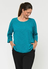 Turkizna teksturirana majica s valovitimi vzorci, okroglo izrezom in rokavi do tri četrtine z gumbi, v kombinaciji z črnimi hlačami.