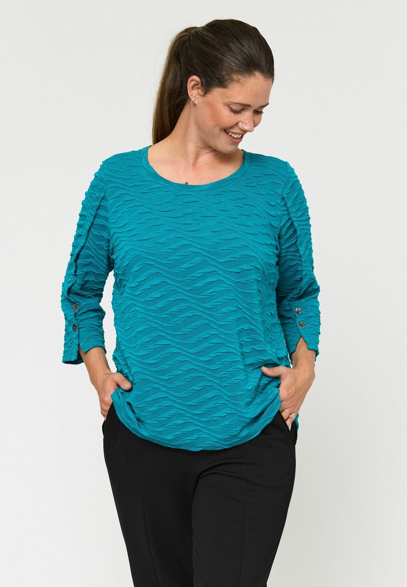 Turkizna teksturirana majica s valovitimi vzorci, okroglo izrezom in rokavi do tri četrtine z gumbi, v kombinaciji z črnimi hlačami.