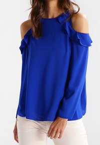 Blå off-shoulder topp med volangdetaljer, långa ärmar och en slät textur. Stylandes med ljusa, figurnära byxor.