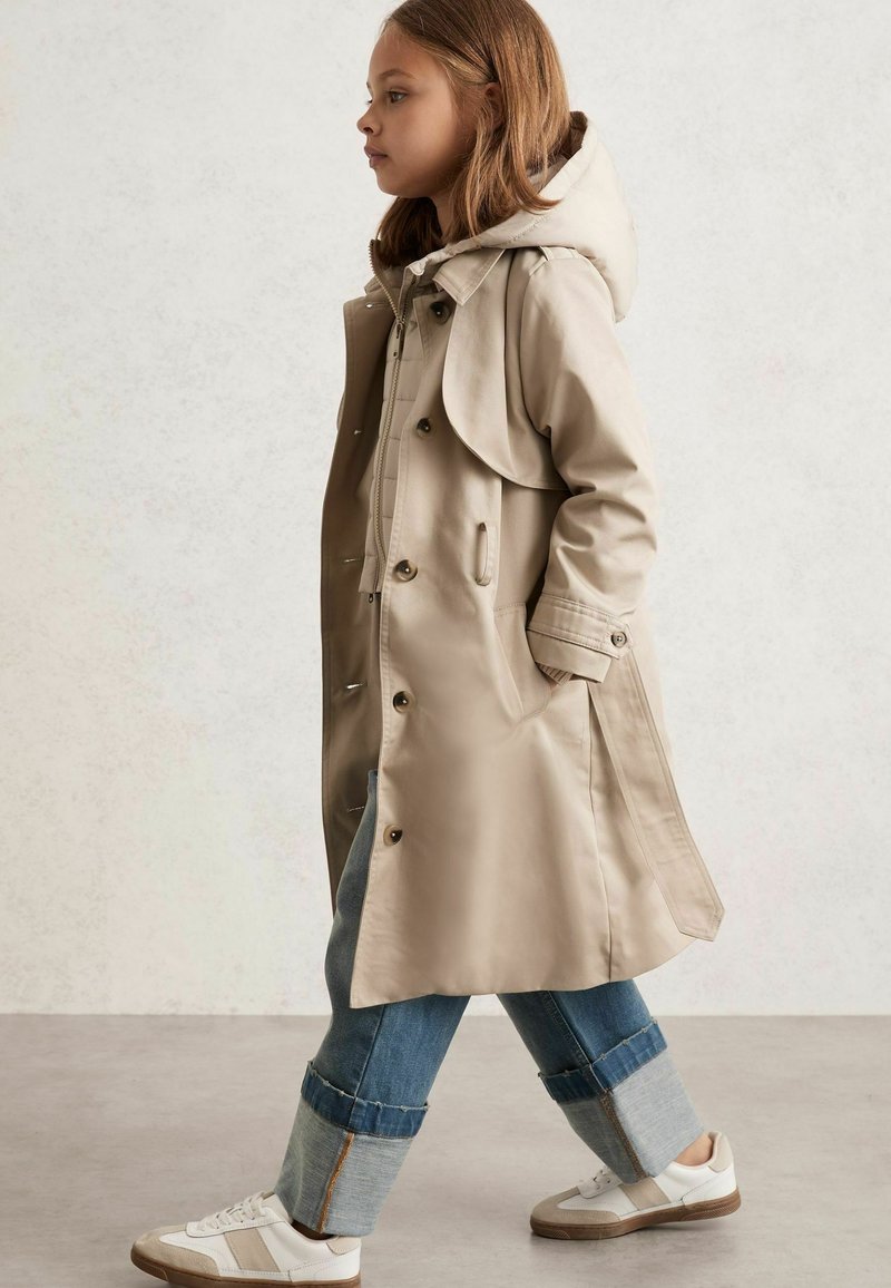 Beige trenchcoat med hætte, knaplukning og sidelommer, båret over blå jeans med optrillede kanter, parret med hvide sneakers.