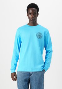 Light blue long-sleeve shirt with a rounded neckline and ribbed cuffs, featuring a circular black graphic on the left chest.