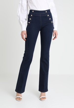 Vrouw gekleed in donkerblauwe high-waisted jeans met zilveren knopen en witte haksandalen, gecombineerd met een witte blouse met lange mouwen.