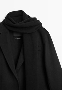 Cappotto di lana nero con ampie rever, abbinato a una sciarpa morbida dello stesso colore drappata attorno al colletto su uno sfondo bianco.