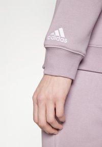 Hand avslappnad vid sidan iförd en lavendel Adidas sweatshirt och matchande byxor, med vit Adidas-logga på ärmslutet.