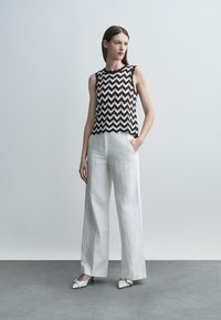 Γυναίκα όρθια με τα χέρια στις τσέπες, φοράει αμάνικο τοπ με μαύρο και λευκό ζιγκ-ζαγκ, λευκό παντελόνι φαρδύ στο κάτω μέρος και λευκές μυτερές γόβες.