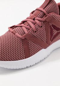 Sportovní bota růžové barvy s texturovaným síťovaným svrškem, plochými tkaničkami a bílou gumovou podrážkou. Obsahuje syntetické prvky pro stabilitu.