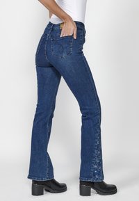 Femme portant un jean en denim bleu foncé avec une broderie florale sur le bas de la jambe et des bottes noires à talons, main dans la poche arrière.
