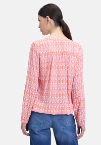Blouse à manches longues rose avec un motif géométrique. Tissu doux, col rond, détail froncé au dos, portée avec un jean bleu.