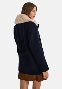 Manteau en laine marine avec un col en fausse fourrure, coupe cintrée, deux poches avant et une ceinture à nouer à la taille. Texture lisse avec des épaules structurées.