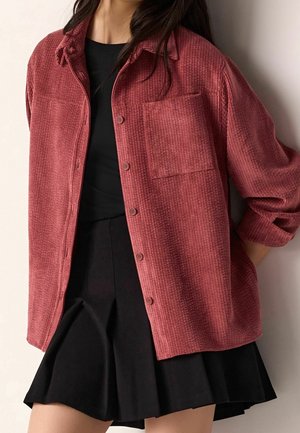 Femme portant une chemise en velours côtelé rouge à manches longues boutonnée sur un top noir et une jupe noire plissée.