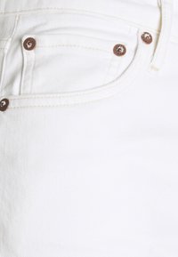 Gros plan sur une poche en denim blanc avec des rivets métalliques bruns visibles et des coutures beiges sur le tissu.