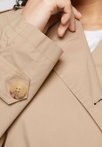 Tessuto di un trench coat beige con una texture liscia. I dettagli includono un grande bottone tondo, color ambra, sulla manica e cuciture a vista.