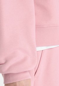 Sweatshirt rose clair mettant en valeur un tissu doux et texturé avec des poignets côtelés et une coupe décontractée, présentant une couche blanche visible en dessous.