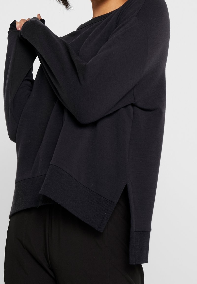 Personne portant un pull noir à manches longues avec des ouvertures pour les pouces et une fente latérale, levant un bras pour montrer le détail de la manche.