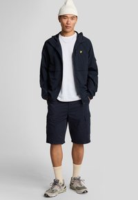 Marine windbreaker met capuchon, geel logo, witte T-shirt, marine cargoshorts, crèmekleurige sokken en grijze sneakers met camouflage-accenten.