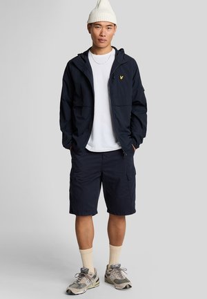 Marinblå vindjacka med huva, gul logotyp, vit t-shirt, marinblå cargoshorts, krämfärgade strumpor och grå sneakers med kamouflage-accenter.