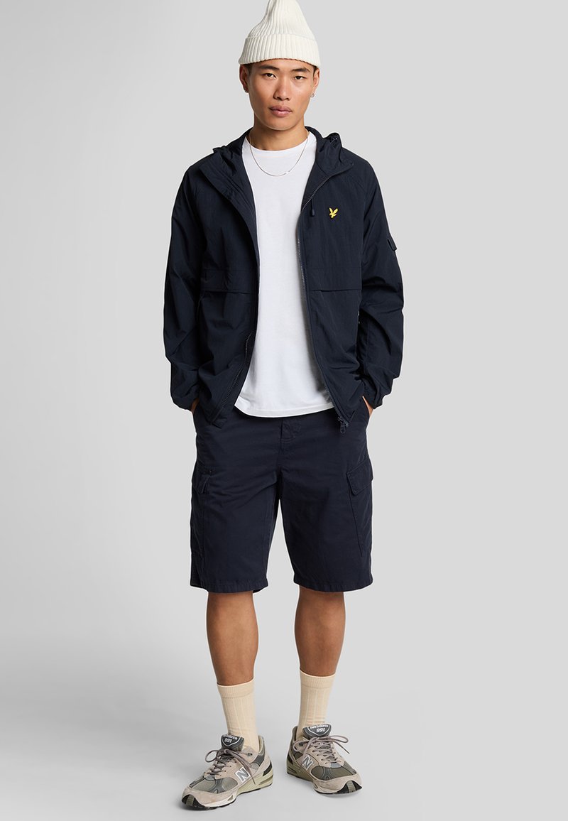 Marine windbreaker met capuchon, geel logo, witte T-shirt, marine cargoshorts, crèmekleurige sokken en grijze sneakers met camouflage-accenten.