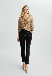 Blusa in stampa animalier color sabbia con accenti neri, abbinata a pantaloni jogger neri con zip laterali e finiture dorate. Tacchi dorati completano l'outfit.