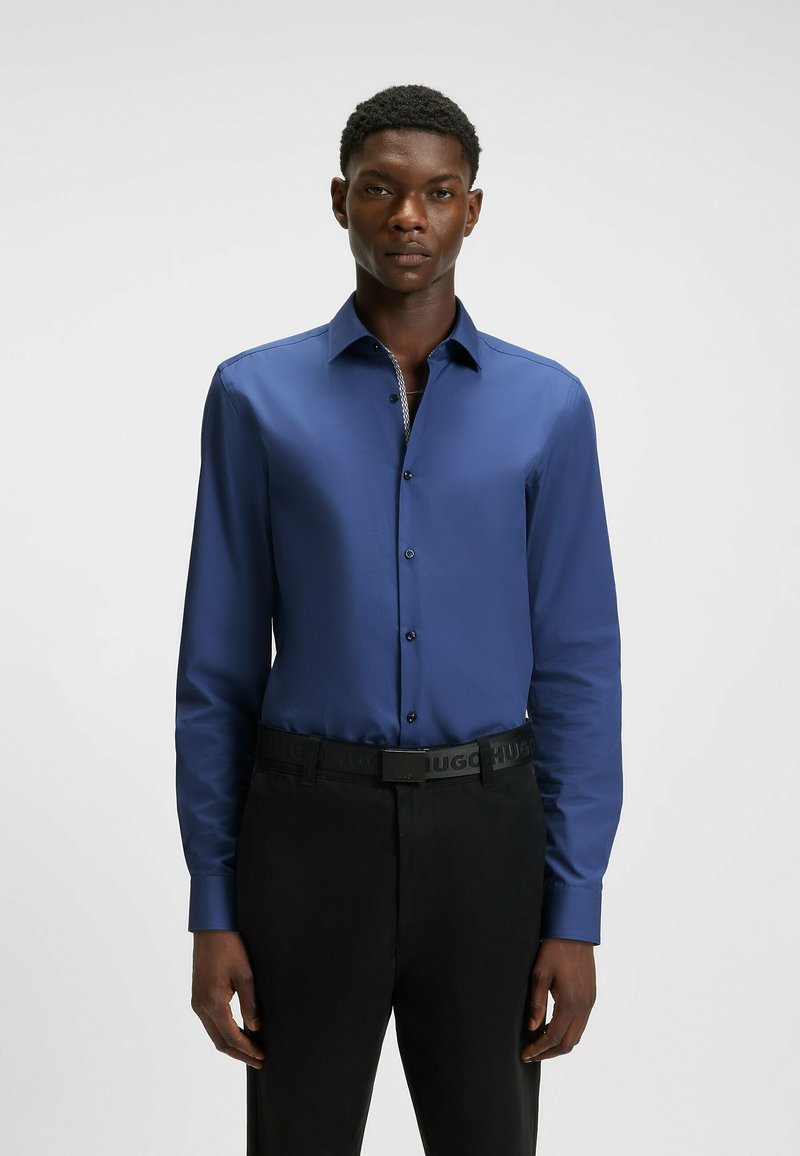Camicia blu a maniche lunghe con una texture liscia e chiusura a bottoni. Indossata con pantaloni neri e una cintura con logo. Design semplice e attillato.