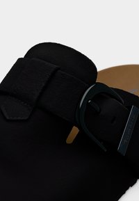 Gros plan sur une bride de sandale en daim noir avec une boucle métallique noire arrondie et une semelle intérieure beige partiellement visible en dessous.
