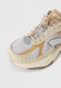 Beige und grauer Sportschuh mit einem strukturierten Mesh-Obermaterial, Gummisohle und kontrastierenden Designelementen. Schnürsenkel mit einem Zugverschluss zur Anpassung.