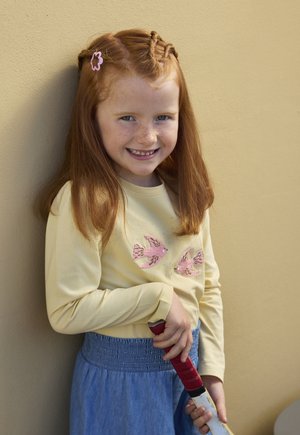 Glimlachend roodharig meisje met gevlochten haar en een roze haarspeld leunt tegen een beige muur, draagt een geel shirt met roze vogels en een blauwe rok, en houdt een hockey stick vast.