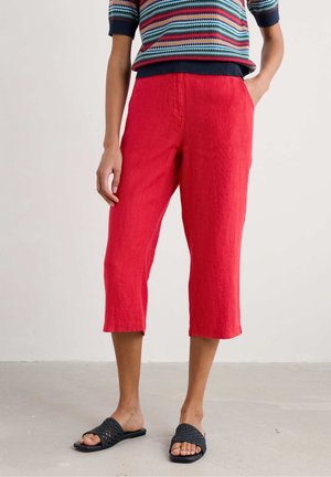 Personne portant un pantalon rouge raccourci avec des poches latérales, un haut coloré à rayures manches courtes et des sandales noires tissées, debout sur un sol gris.