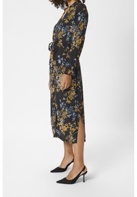 Abito nero con motivo floreale blu e giallo, maniche lunghe, vita legata, spacco laterale; abbinato a tacchi slingback neri con punta affilata.