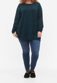 Ciemny zielono-niebieski, oversize'owy sweter z długimi rękawami i prostym wykończeniem, w zestawie z niebieskimi skinny jeansami i czarnymi butami wsuwanymi z złotymi akcentami.