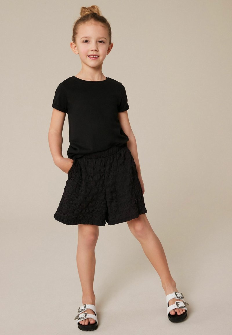 Camiseta negra de manga corta combinada con pantalones cortos negros texturizados y sandalias blancas con hebillas plateadas. Diseño sencillo, corte casual.