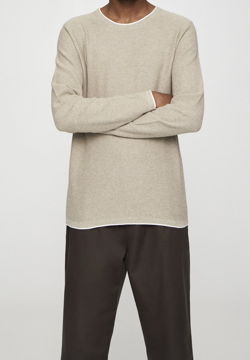 Pull texturé beige clair à manches longues, col rond, avec un détail blanc au col et à l'ourlet. Associé à un pantalon foncé.