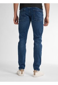 Jeans ajustados de color azul oscuro con una cintura de altura media, con bolsillos traseros y un acabado suave y texturizado. Combinados con zapatos blancos y una camisa negra.