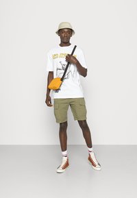 T-shirt graphique blanche avec texte jaune, shorts cargo verts olive, sac bandoulière jaune, chapeau seau beige et baskets montantes marron.