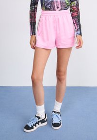Shorts de sport roses avec une taille élastique et des rayures sur les côtés. Portés avec des baskets noires avec des lacets bleu clair et des chaussettes blanches jusqu'aux chevilles.