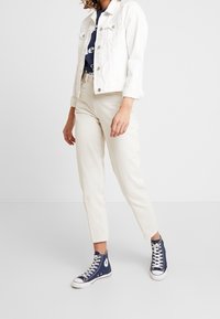 White denim jacket, navy graphic t-shirt, cream tapered trousers, and blue high-top sneakers. Focus on material and color contrasts.