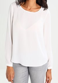 Blouse blanche à manches longues en tissu transparent avec un col rond et un ourlet légèrement asymétrique ; portée avec un pantalon gris clair. Texture lisse et légère.