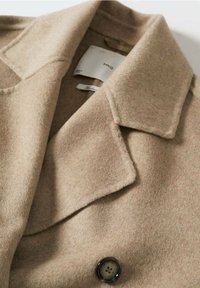 Beige wollen jas met een brede revers, voorzien van een zwart knoopdetail. Zachte textuur met een strak, minimalistisch ontwerp en zichtbare hangtags.