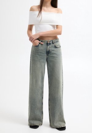 Vrouw die een witte off-shoulder cropped top draagt en een high-waisted wijde grijze jeans met vervaagde look, staand met gekruiste handen.