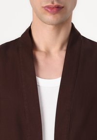 Brun bomullsjakke med sjalkrage, vist over en hvit ribbestrikket skjorte. Fokuser på utringningen og tekstur i stoffet.