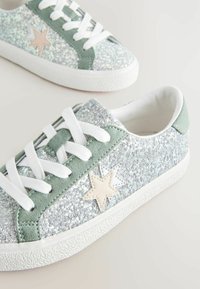 Glitzernde Sneakers mit einem silbernen Obermaterial, grünen Velourslederakzenten, einer strukturierten weißen Sohle und weißen Schnürsenkeln. Mit einem beigen Sternenmotiv an der Seite.