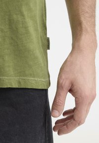 Oliwkowa bawełniana koszulka o prostym kroju, z okrągłym dekoltem i widocznym przeszyciem na dole. Czarna metka po boku.