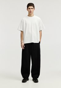 Tricou alb din bumbac cu croi lejer, mâneci scurte și decolteu rotund, purtat cu pantaloni negri largi și adidași negri.