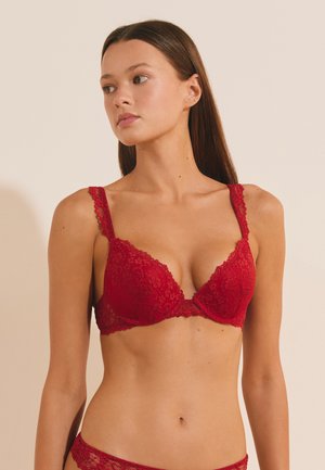 Femme aux longs cheveux bruns portant un soutien-gorge en dentelle rouge et une lingerie assortie, regardant légèrement vers la gauche sur un fond beige uni.