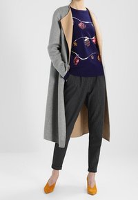 Abrigo de lana gris con forro interior color beige, suéter azul marino con patrones ornamentales coloridos, pantalones negros a rayas y tacones amarillos.