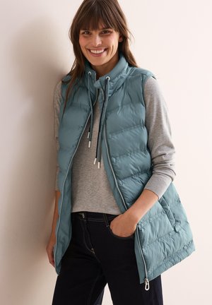 Gilet matelassé bleu-vert clair avec col montant, fermeture éclair à l'avant et cordon de serrage. Porté sur un pull gris, associé à un jean foncé. Matière lisse et isolante.